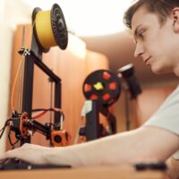 Mann sitzt hinter einem Labortisch und stellt einen 3D-Drucker ein