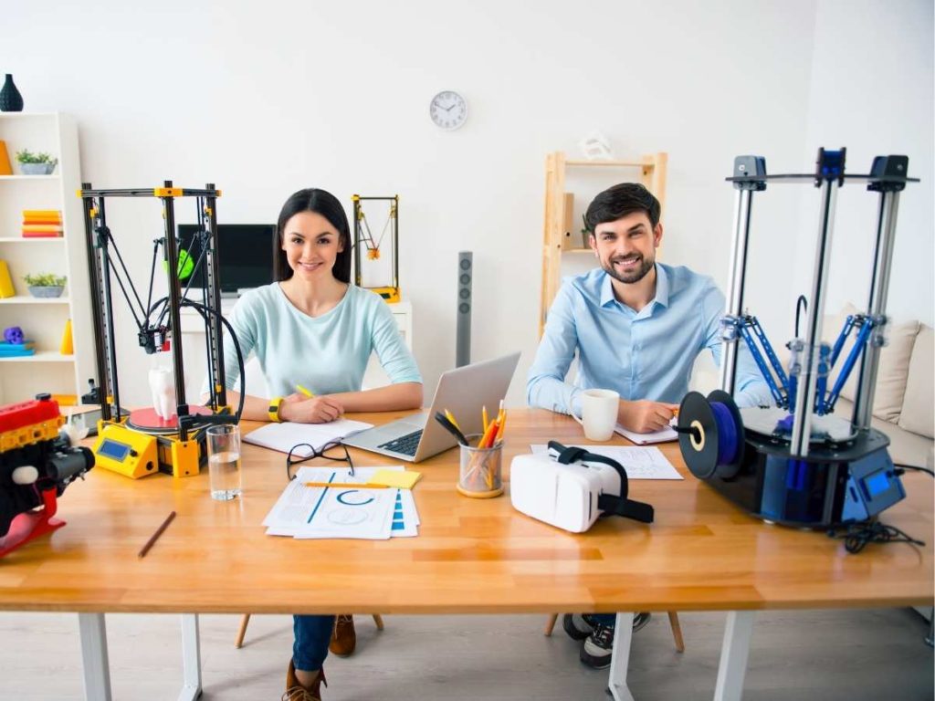 2 persons sitting next to 2 3d printers comparing them