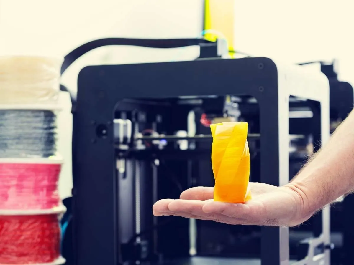 3d printer in the background with a 3d printed object holding in hand