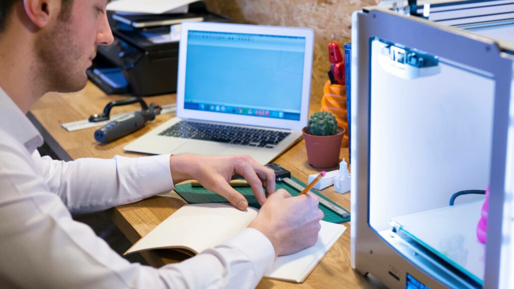 3d printing an object in 3d printer with a labtop on the desk and person sitting next to it