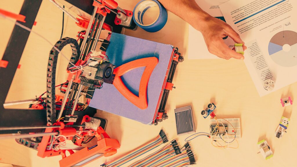 A 3D printer creating an orange object, surrounded by electronics and a person's hand.