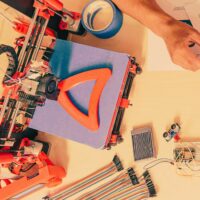A 3D printer creating an orange object, surrounded by electronics and a person's hand.